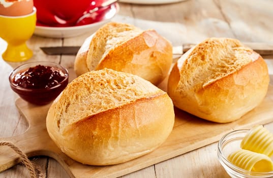 Three whole buns of freshly baked white bread served with jam and butter on wooden cutting board in restaurant
