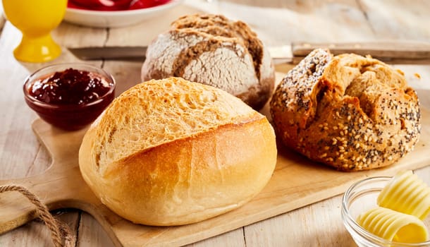 Whole buns of various kinds of fresh baked bread served in restaurant on wooden cutting board, with butter and jam in glass bowls