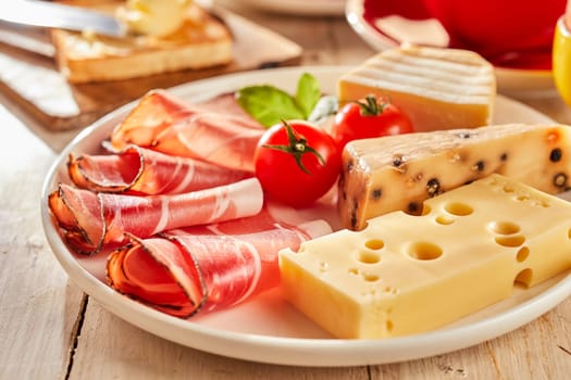 Variety of cheese served with sliced bacon and fresh tomatoes on white plate in restaurant. Viewed in closeup from the side