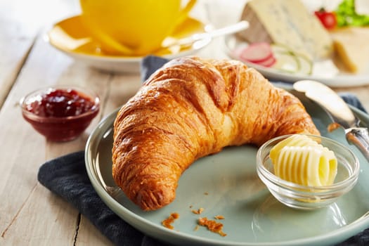 Whole crunchy fresh croissant served on ceramic plate with butter curls in glass bowls. Restaurant table in close-up