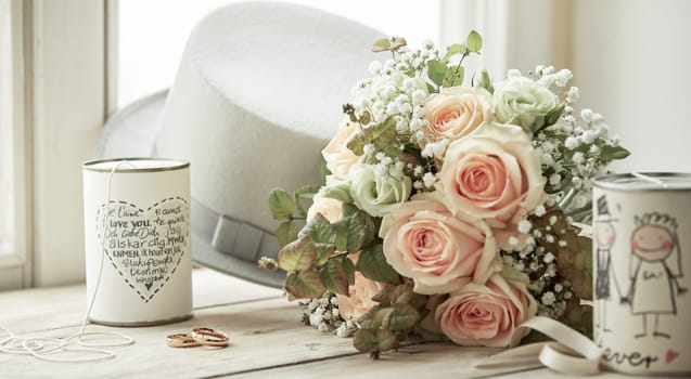 Bright composition for marriage day with wedding rings, bridal bouquet of pink roses, brooms white hat and cans with decorations and wishes, sitting on bright window sill