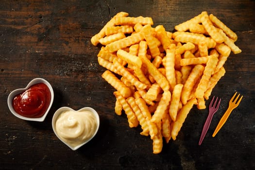 Crinkle cut crispy deep fried Pommes Frites arranged in a heart shape on wood with matching bowls of ketchup and mayonnaise viewed from above