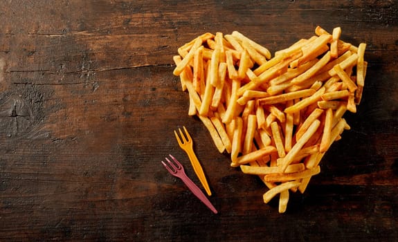 Heart shaped still life of French fries, pommes frites or potato chips viewed from above on vintage wood with copy space and plastic forks