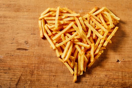 Close-up view from above of a heart shape made of tasty French fries served on a wooden rustic table as concept for love or fast food