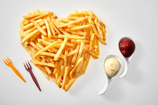 Heart shaped arrangement of French fries with tomato sauce and mayonnaise in ceramic spoons and plastic forks on a white background