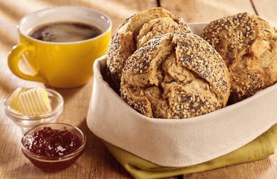 Fresh baked buns of brown wholewheat bread served for breakfast in fabric bag, with bowl of jam, curls of butter and cup of coffee, on wooden table and viewed in close-up