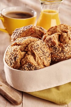 Fresh baked dark bread buns with seeds, served in fabric bag with juice and coffee in background on wooden table