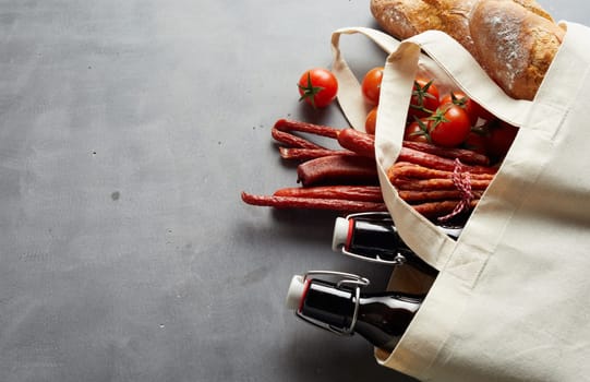 Craft beer, spicy sausage and baguettes in an eco friendly reusable fabric shopping bag over slate with copy space