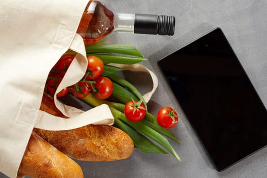 Tablet with fresh groceries and bottle of wine in a reusable textile carrier bag to save on pollution from plastics in the environment