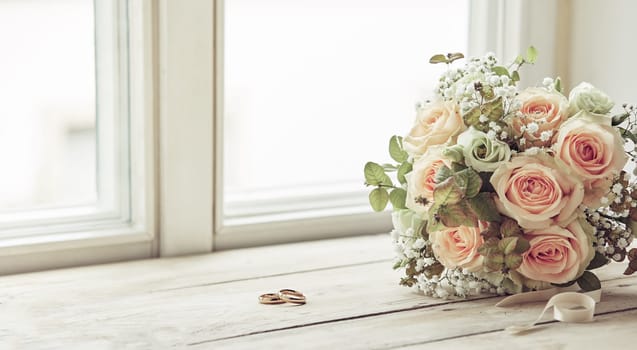 Pair of wedding rings and bridal bouquet of pink roses, sitting on bright window sill, before the ceremony