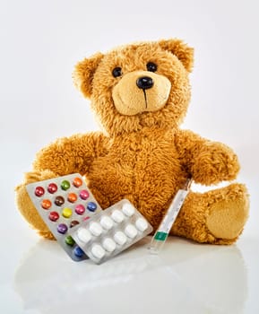 Toy bear with medicine, thermometer and colorful pills in blister pack, sitting on glossy surface against white background. Viewed from its front. Pediatrician office and children health concept