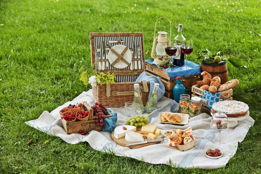 Gourmet picnic spread with fresh fruit, cheese, wraps, crusty bread, wine and dessert surrounding a wicker hamper on green grass in a park