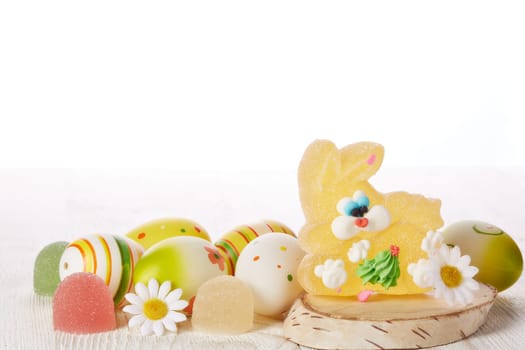 Still life with colored easter eggs and easter rabbit from sweet fruit jelly lying on a light wooden table on white background. Greeting card.
