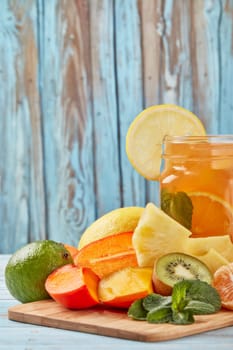 Fruit lemonade with ice, leaves of mint, lemon, pineapple, mango and lime slices standing on blue wooden table. Homemade lemonade with mix fruits. Healthy cool beverage, fruit juice