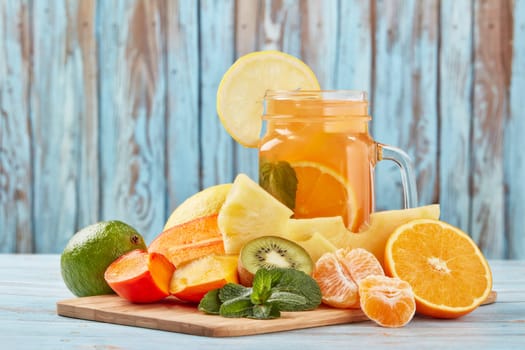Fruit lemonade with ice, leaves of mint, lemon, pineapple, mango and lime slices standing on blue wooden table. Homemade lemonade with mix fruits. Healthy cool beverage, fruit juice