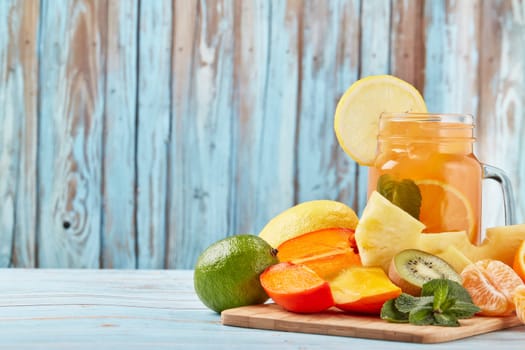 Fruit lemonade with ice, leaves of mint, lemon, pineapple, mango and lime slices standing on blue wooden table. Homemade lemonade with mix fruits. Healthy cool beverage, fruit juice