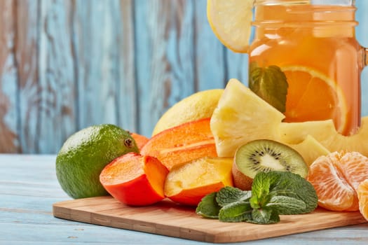 Fruit lemonade with ice, leaves of mint, lemon, pineapple, mango and lime slices standing on blue wooden table. Homemade lemonade with mix fruits. Healthy cool beverage, fruit juice