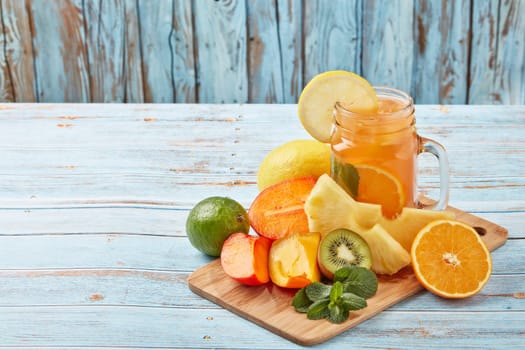 Fruit lemonade with ice, leaves of mint, lemon, pineapple, mango and lime slices standing on blue wooden table. Homemade lemonade with mix fruits. Healthy cool beverage, fruit juice