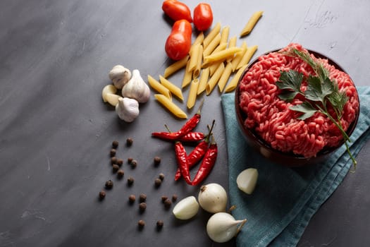 Still life with fresh forcemeat from beef in a bowl, vegetables, pasta and spices on a dark background. Top view