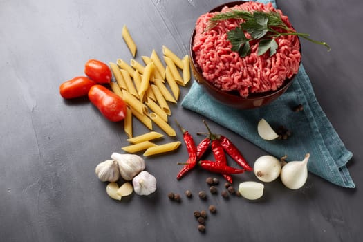Still life with fresh forcemeat from beef in a bowl, vegetables, pasta and spices on a dark background. Top view