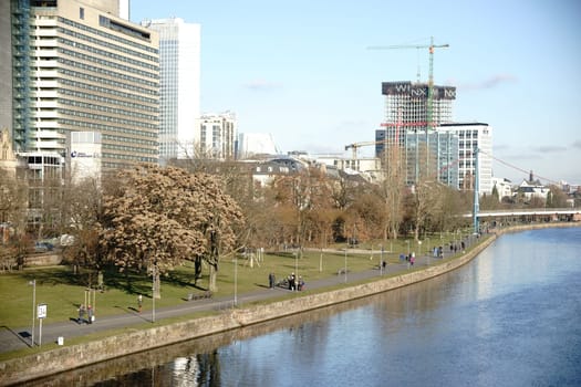 Frankfurt, Germany - January 05, 2017: Strollers walk along the waterfront along the Main river with the skyline of the Frankfurt financial district behind it on January 05, 2017 in Frankfurt..