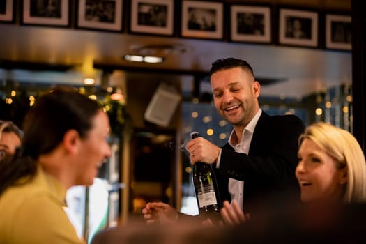 Mid adult waiter opening a bottle of prosecco for two women as they sit in a restaurant.