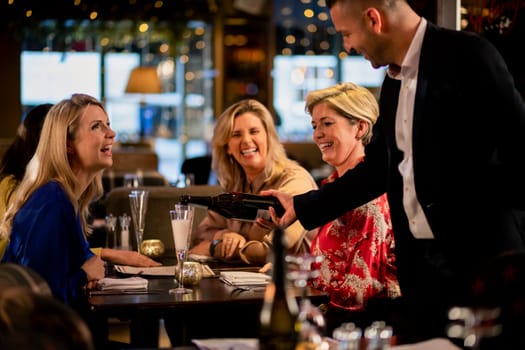 Mature waiter pouring a glass of prosecco for a table with three mature women sitting, laughing and smiling.