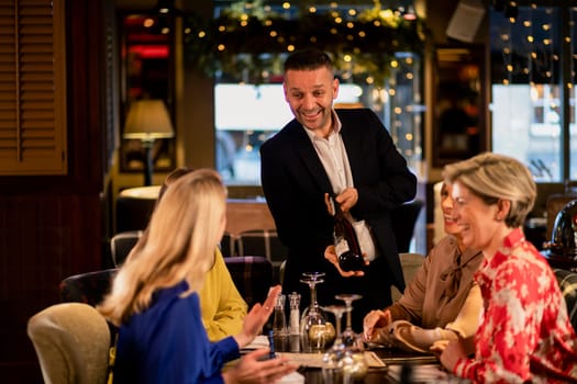 Mature waiter reccomending a bottle of champagne to a small group of mature women. They are sitting at a table in a restaurant.