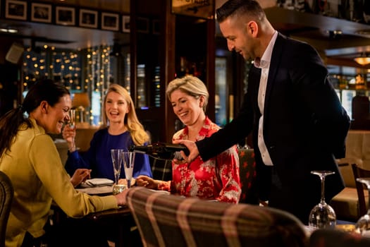 Mature waiter pouring a glass of prosecco for a table with three mature women sitting, laughing and smiling.