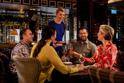 Two couples sitting at a table in a restaurant. The waitress is pouring a glass of while while a mature female holds the glass.