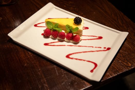 Slice of lemon cheesecake on a plate. It has been garnsihed with a blackberry and raspberries.