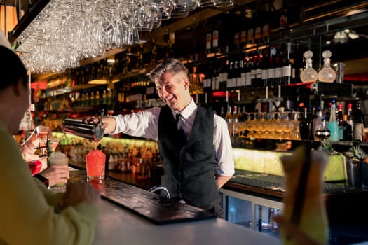 Young bartender pouring a cocktail out of a coktail shaker at the bar counter.