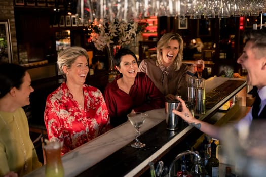 Small group of mature women enjoying a night, sitting at the bar. They are laughing with the young bartender who is making a cocktail.
