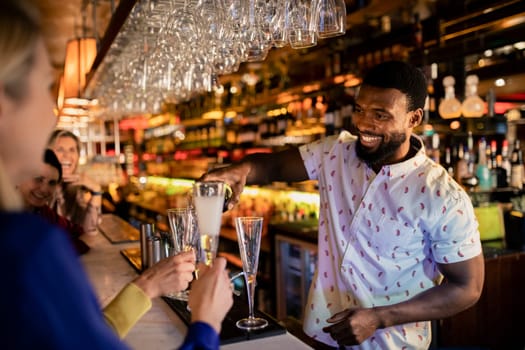 Bartender filling up a mature womans glass with prosecco. She is sitting at the bar counter with her friends.