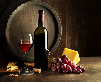 Bottle of red wine with glass and cheese on wooden and barrel background.