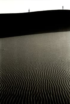 Black and white view of two young women standing high on sand dune of the desert, the first is in contemplation while the other is approaching her walking, Merzouga, Morocco