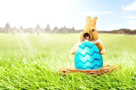 Little bunny holding colorful easter egg in the nest on the grass field with trees and blue sky background. Happy Easter