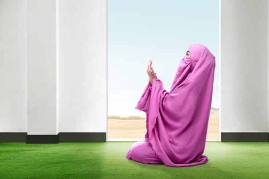 Beautiful asian woman in pink veil sitting in praying position and raise the hands on the carpet inside the room with open door and blue sky on the background