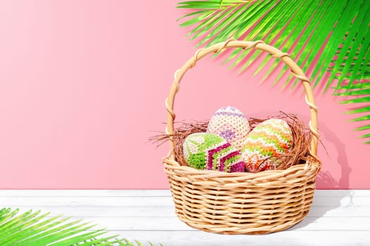 Colorful easter eggs on the nest in wooden basket on white wooden table with palm leaves and pink wall background. Happy Easter