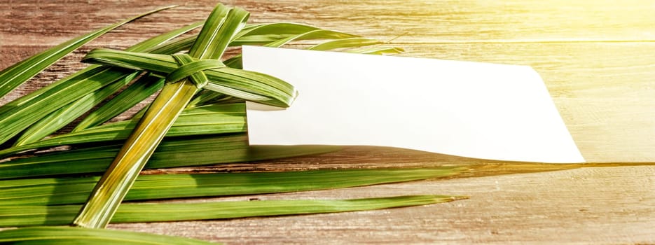 Closeup view of cross shape of palm leaf and palm branches with white blank paper and ray in wooden background. Palm Sunday concept