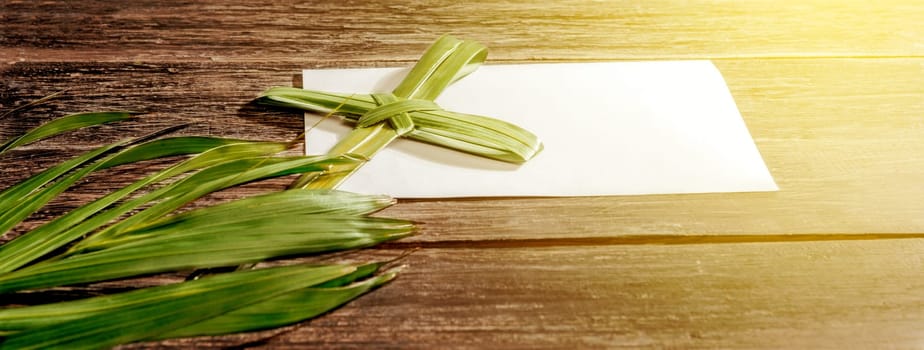 Closeup view of cross shape of palm leaf and palm branches with white blank paper and ray in wooden background. Palm Sunday concept