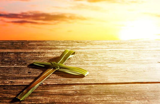 Cross shape of palm leaf on wooden table with rays of sunset background. Palm Sunday concept