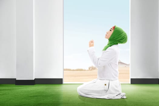 Pretty asian woman in veil sitting in praying position on the carpet rise the hands and looking up inside the room with open door and blue sky background