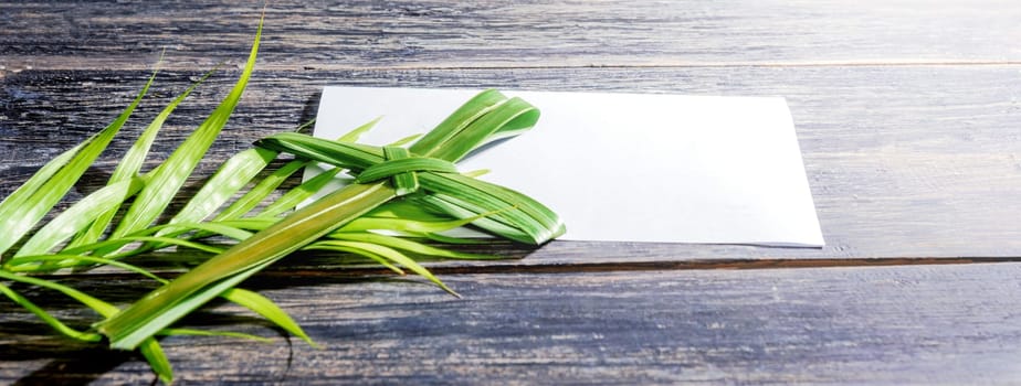 Closeup view of cross shape of palm leaf and palm branches with white blank paper on wooden background. Palm Sunday concept