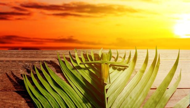 Cross shape of palm leaf and palm branches on wooden table with rays of sunset background. Palm Sunday concept