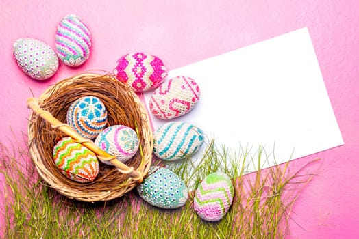 Colorful easter eggs in the nest and grass with blank white paper on the pink background. Happy Easter