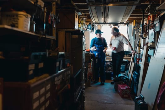 Two senior men are in a construction workshop. One man is explaining something and the other man is drinking a cup of tea while listening.