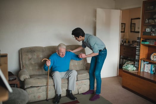 Teenage boy is helping his grandfather stand up from the sofa in his living room.