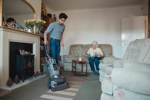 Teenage boy is hoovering his grandmother's living room for her while she reads a newspaper.