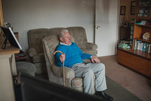 Senior man is sitting in his living room at home with a walking stick.